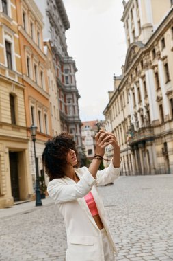 A woman in a white blazer and pink top takes a selfie in a picturesque European street. clipart