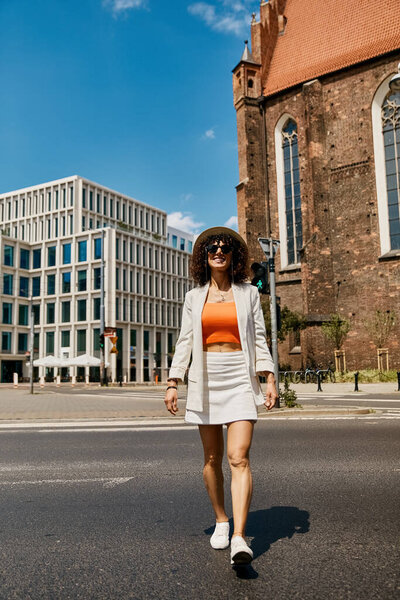 Стильная женщина в белом пиджаке и юбке ходит по улице в Европе.