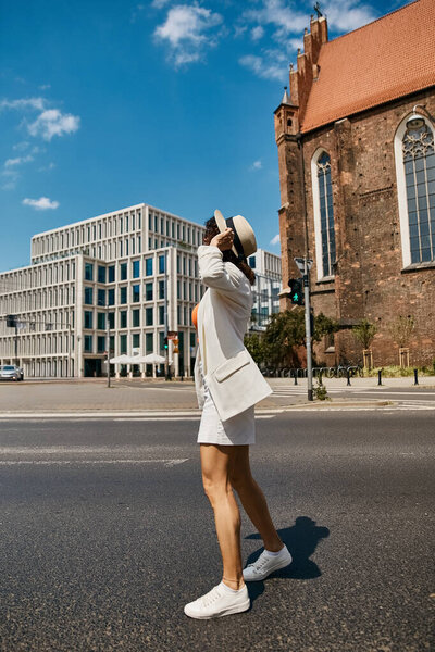 Женщина в белом пиджаке и юбке идет по улице в европейском городе, наслаждаясь солнцем.