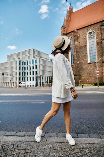 A woman in a white blazer and straw hat walks down a cobblestone street in a European city.