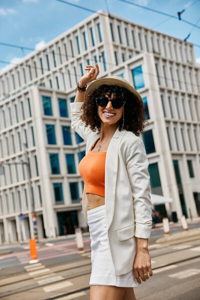 Стильная женщина прогуливается по городской улице Европы в белом блейзере и оранжевом топе.