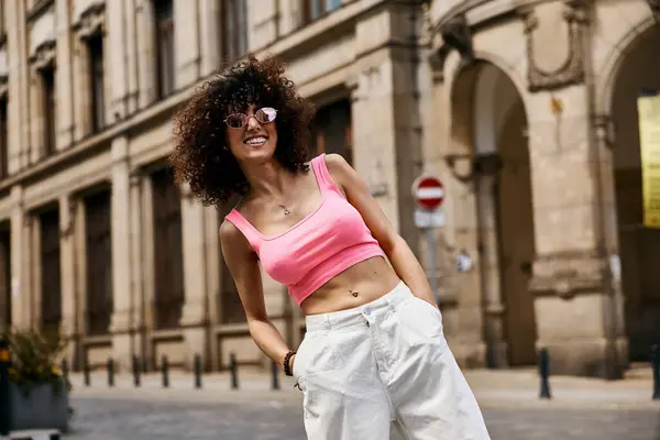 stock image A woman in a pink crop top and white pants smiles while exploring the streets of a European city.