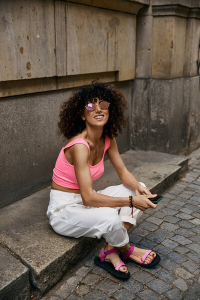 A fashionable woman in a pink tank top and white pants sits on a step in a European city.
