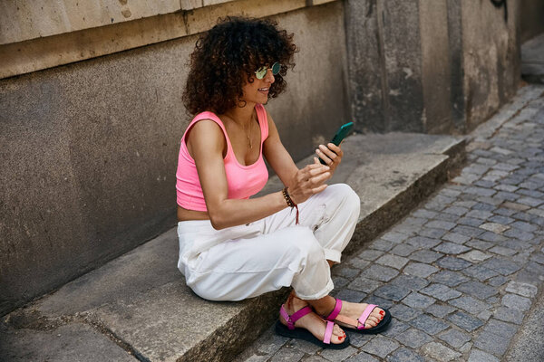 A woman wearing stylish attire sits on a step in a European city, checking her phone.