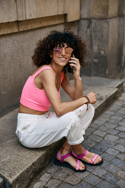 A stylish woman wearing a pink crop top and white pants talks on her phone while exploring cobblestone streets in Europe.