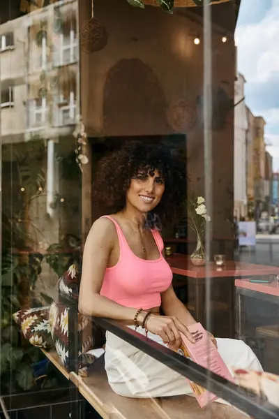 stock image A beautiful woman in a pink tank top and white pants sits outside a cafe in Europe, looking out the window.