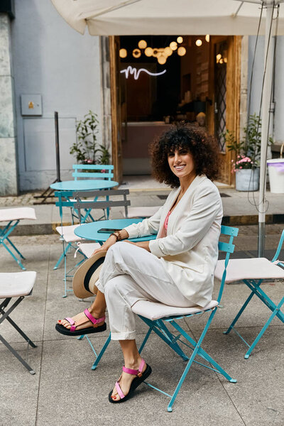 A stylish woman in white pants and a blazer enjoys a European cafe setting.