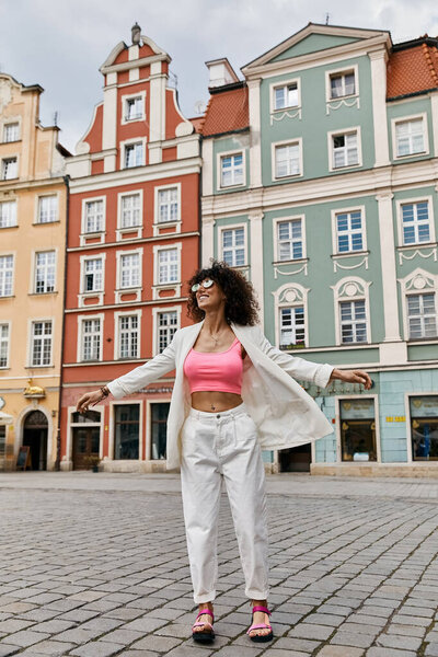 Стильная женщина в белом блейзере и белых брюках позирует перед красочными зданиями в Европе.