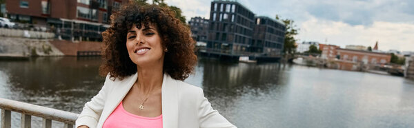 Красивая женщина в стильном белом блейзере и розовом топе наслаждается солнечным днем в своем европейском путешествии, стоя у канала.