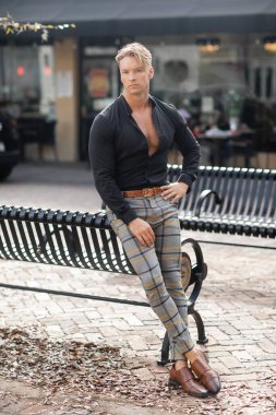 Siyah gömlekli ve ekose pantolonlu yakışıklı sarışın bir adam Orlando, Florida 'da bir bankta poz veriyor..