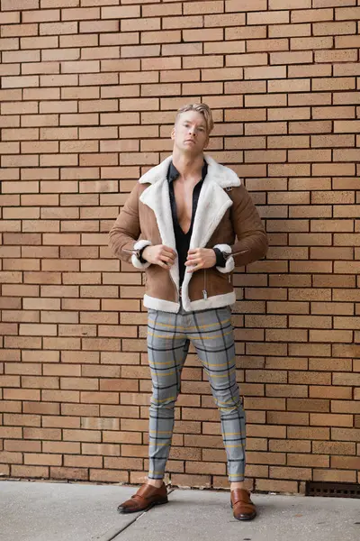 stock image A handsome blonde man wearing a brown jacket and plaid pants poses in front of a brick wall