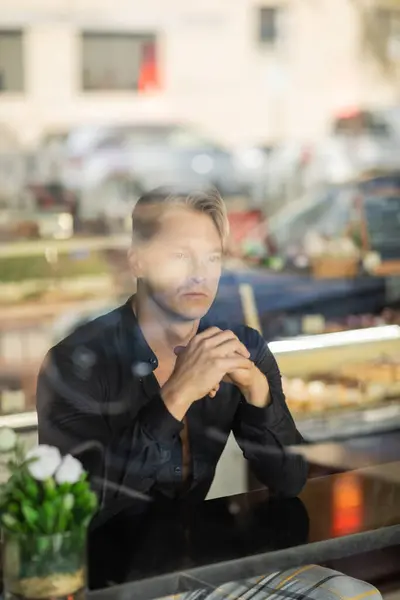 stock image A blonde man, dressed in a stylish black shirt, sits thoughtfully at a table in cafe.