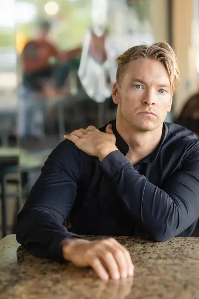 stock image A handsome blonde man in stylish attire sits at a table, thoughtfully looking off to the side, on a street in Orlando, Florida.