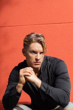 Yakışıklı sarışın bir adam, şık giyinmiş, düşünceli bir şekilde kırmızı duvarın yanında oturuyor.