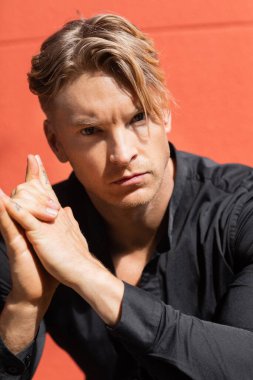 Şık giyinmiş yakışıklı sarışın bir adam kırmızı duvarlı bir sokakta duruyor.