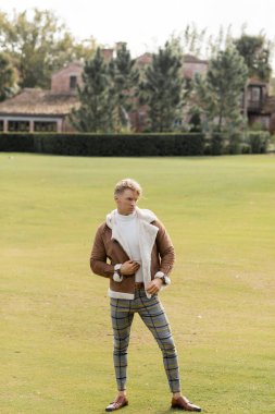 A handsome blonde man in stylish attire walks through a lush green park in Miami. clipart