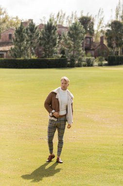 A stylish blonde man in a brown jacket and plaid pants walks across a green park in Miami. clipart