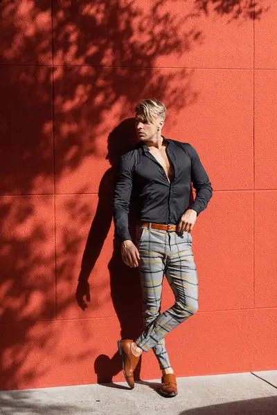 stock image A handsome blonde man in a black shirt and plaid pants leans against a red wall in Orlando, Florida.