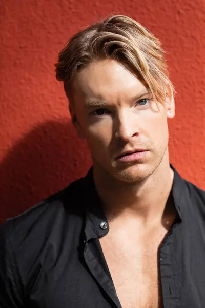 stock image A handsome blonde man in stylish attire stands on a street with red wall