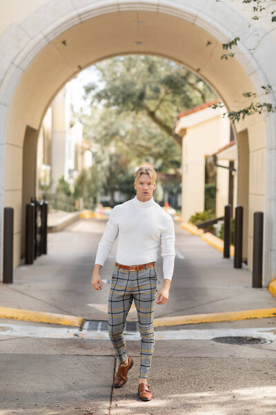 A blonde man wearing a white turtleneck and plaid pants poses on a street in Florida.