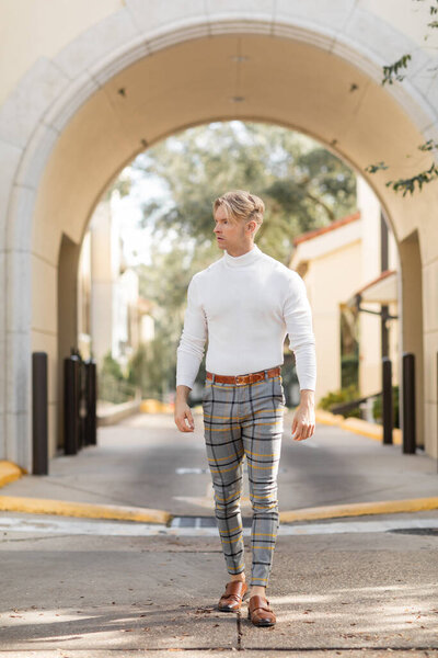 A blonde man in a white turtleneck and checkered pants poses on a street in Florida.