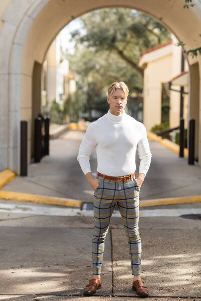 A blonde man wearing a white turtleneck and plaid pants poses with hands in his pockets under an arched entrance in Florida.
