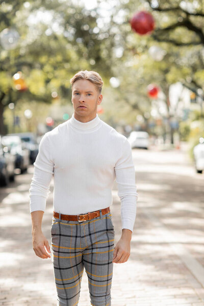 Блондин в белой водолазке и в клетчатых штанах позирует на улице во Флориде.