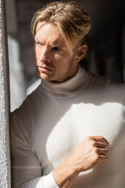 Stock image A blonde man in a white turtleneck stands against a building in Florida, basking in the sunlight.