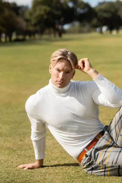 stock image A stylish blonde man in a white turtleneck and plaid pants poses on a grassy park in Miami.