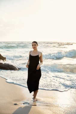 A girl in a black sundress walks Miami sandy beach, ocean waves cresting behind her. clipart