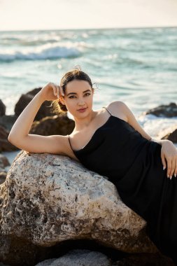 Siyah elbiseli genç bir kadın Miami sahilinde bir kayaya yaslanmış okyanusa bakıyor..