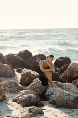 Genç bir kadın Miami sahilinde büyük bir kayanın üzerinde oturuyor, siyah bir elbise giyiyor.