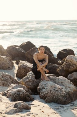 Siyah bir elbise ve hasır şapka giyen genç bir kadın okyanusa tepeden bakan kayalarda oturur ve düşünceli bir şekilde ufka bakar..