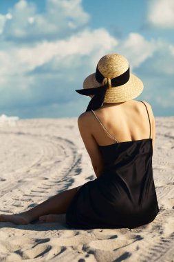 Siyah elbisesi ve hasır şapkası olan genç bir kadın Miami, Florida 'da beyaz kumsalda oturmuş okyanusu seyrediyor..