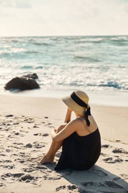 A young woman, wearing a black sundress and straw hat, sits on the sandy beach gazing out at the blue ocean waves. clipart