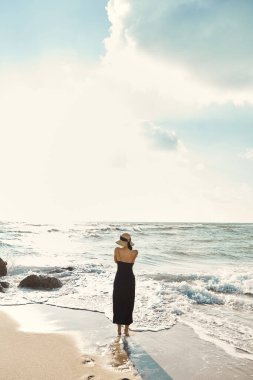 A woman in a black sundress and straw hat stands on the beach, gazing out at the ocean where gentle waves lap at the shore. clipart