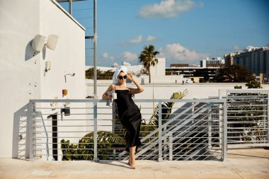 A woman in a black slip dress and sunglasses leans against a railing on a villa rooftop overlooking a Miami pool. clipart