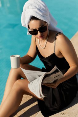 Siyah iç çamaşırlı ve güneş gözlüklü şık bir kadın Miami 'de havuz kenarında rahatlıyor, dergi ve içkinin tadını çıkarıyor..