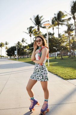 A young woman in sunglasses and a floral skirt skates down a palm tree-lined pathway in Miami. clipart