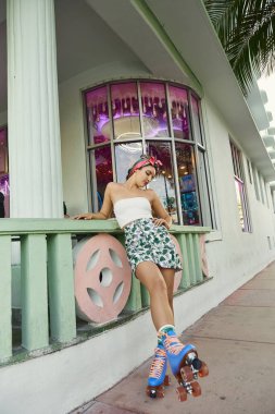 A young woman in sunglasses and a floral skirt skates through the streets of Miami. clipart