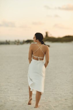 A young woman strolls barefoot along the beach in Miami, her white dress flowing behind her. clipart