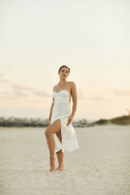 Beyaz elbiseli genç bir kadın Miami 'de kumlu bir sahilde duruyor, batan güneşin sıcacık parıltısıyla yıkanıyor..