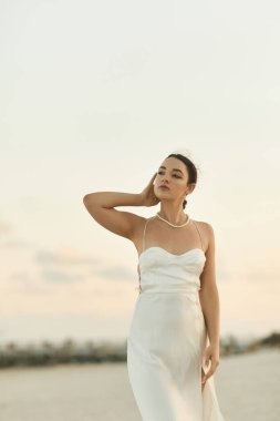 Beyaz elbiseli, saçları arkaya çekilmiş genç bir kadın güneş batarken Miami 'de kumlu bir sahilde duruyor..