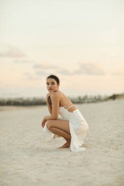 A young woman in a white dress sits on the beach in Miami, looking out at the horizon. clipart