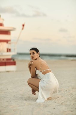 Beyaz elbiseli genç bir kadın Miami sahilinin kumsalında oturuyor, gözleri uzaklara bakıyor..