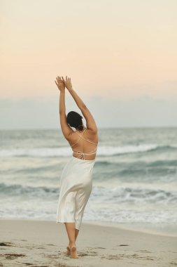 Beyaz elbiseli genç bir kadın kumlu bir Miami sahilinde duruyor, kolları açık, serin akşam melteminin tadını çıkarıyor..