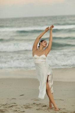 A woman in a white dress with a slit stands on a sandy Miami beach, arms raised, gazing towards the setting sun. clipart