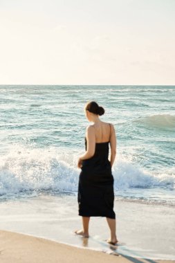 Siyah elbiseli bir kadın Miami Beach, Florida boyunca yürür ve dalgalara bakar..