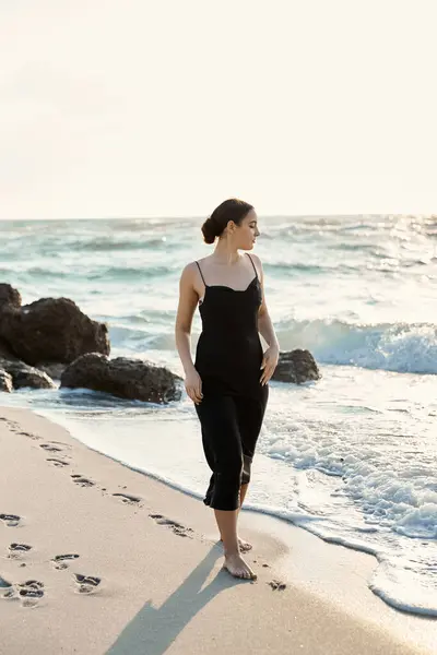 stock image A young woman in a black sundress walks along the shoreline of Miami Beach, leaving footprints in the sand.