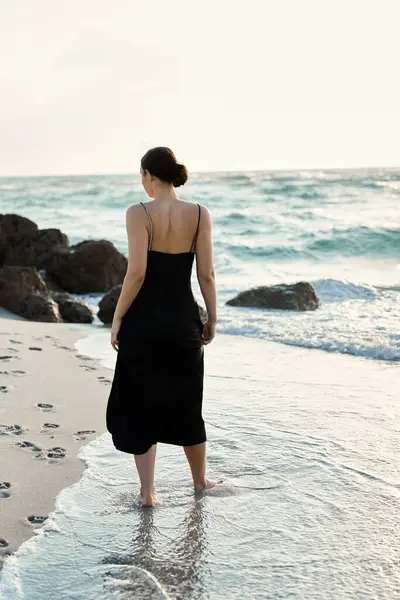 stock image A woman in a black sundress walks along a sandy beach, the water lapping at her feet.